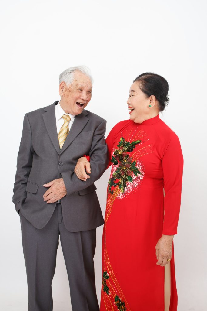 一對東方長者夫妻並肩微笑——丈夫穿灰西裝、妻子穿紅色傳統服飾，象徵子女替父母補拍的那張遲到的婚紗合照